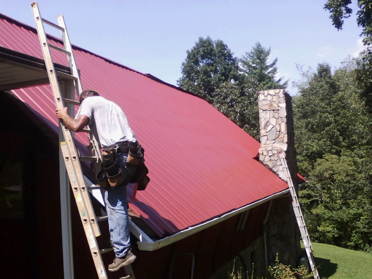 Roofer on a ladder beside a red metal roof and stone chimney — Soffit & Fascia Repair by Roofing Brothers in Macungie